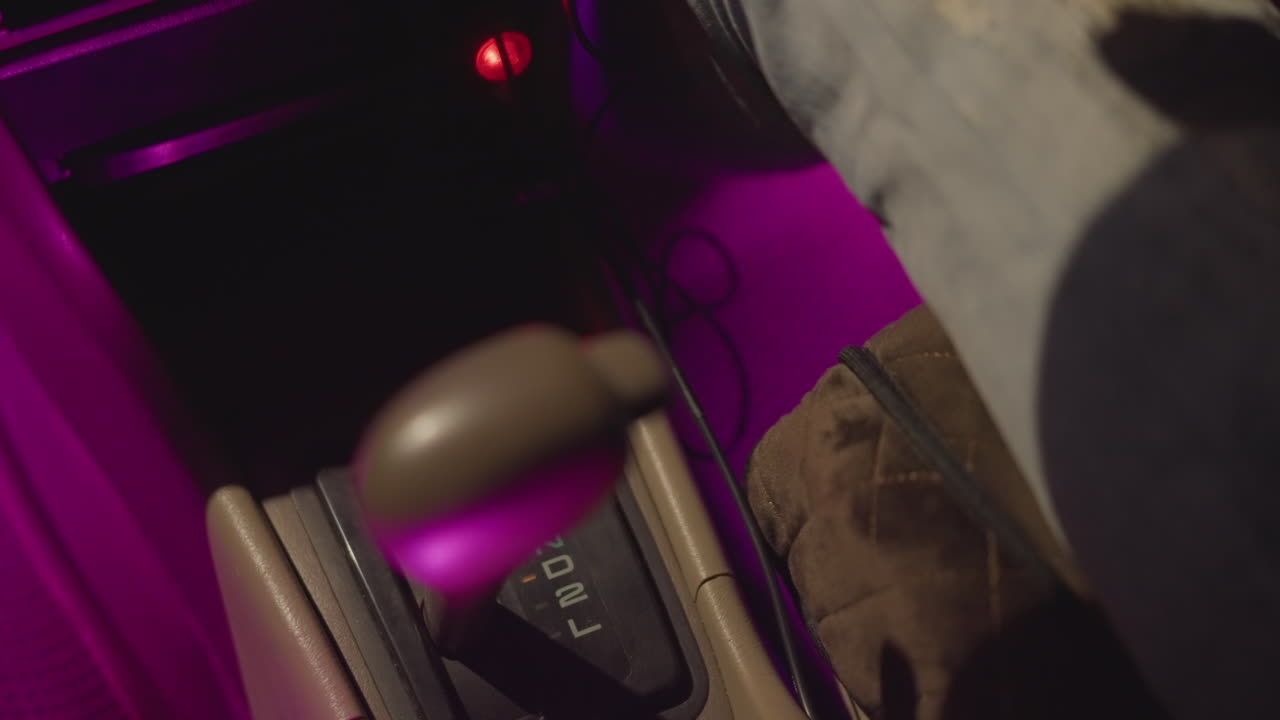 interior de coche nocturno con luces de neón, mano cambiando de marcha bajo una vibrante luz púrpura, primer plano que muestra el reloj, los vaqueros, la bota y la consola, dedos ajustando el mando automático mientras suena la música, ambiente urbano relajado