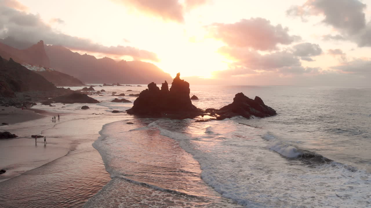 Flying backwards above coastline and rocks with beautiful sunset light at Benijo beach in Tenerife, Canary Islands. Big wave is crashing on rocks and spraying.