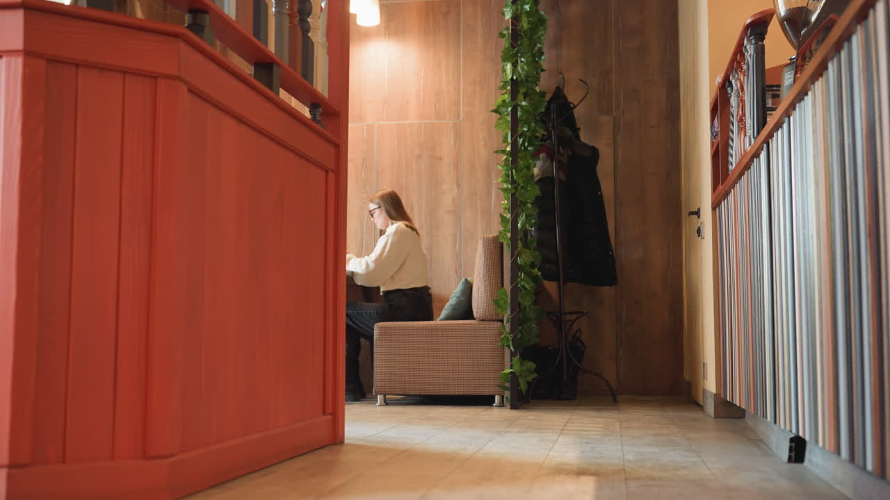 Young woman seated alone under warm hanging light operating phone with calm expression, background includes cloth hanger on wall with minimalistic interior design