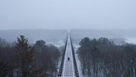 Filmische-Aufsteigende-Luftaufnahme-Eines-Mannes,-Der-In-Einer-Verschneiten-Umgebung-Eine-Sehr-Lange-Eisenbahnbrücke-Entlang-Geht