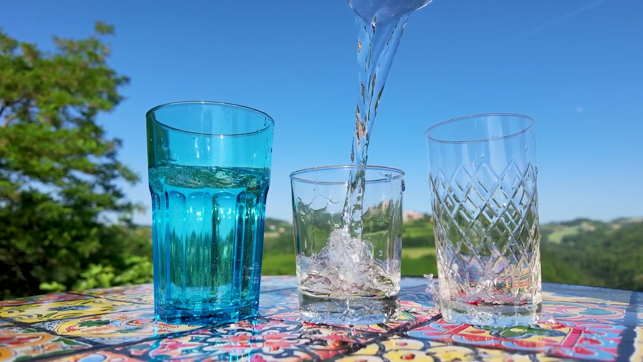 Acqua frizzante si versa in un bicchiere trasparente su un tavolo colorato sotto la luce intensa del sole.