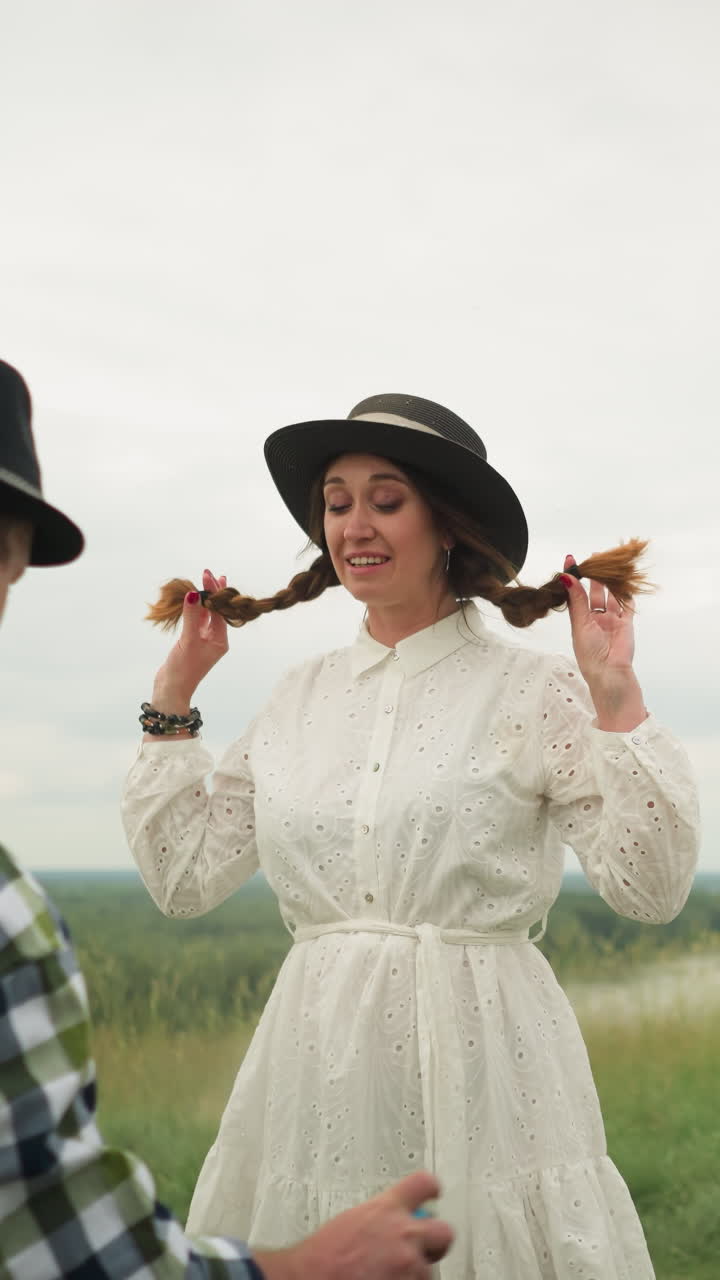 un hombre con una camisa a cuadros pulveriza repelente de mosquitos sobre una mujer con un sombrero elegante y un vestido blanco. están de pie en un campo de hierba exuberante con un lienzo parcialmente pintado en el fondo