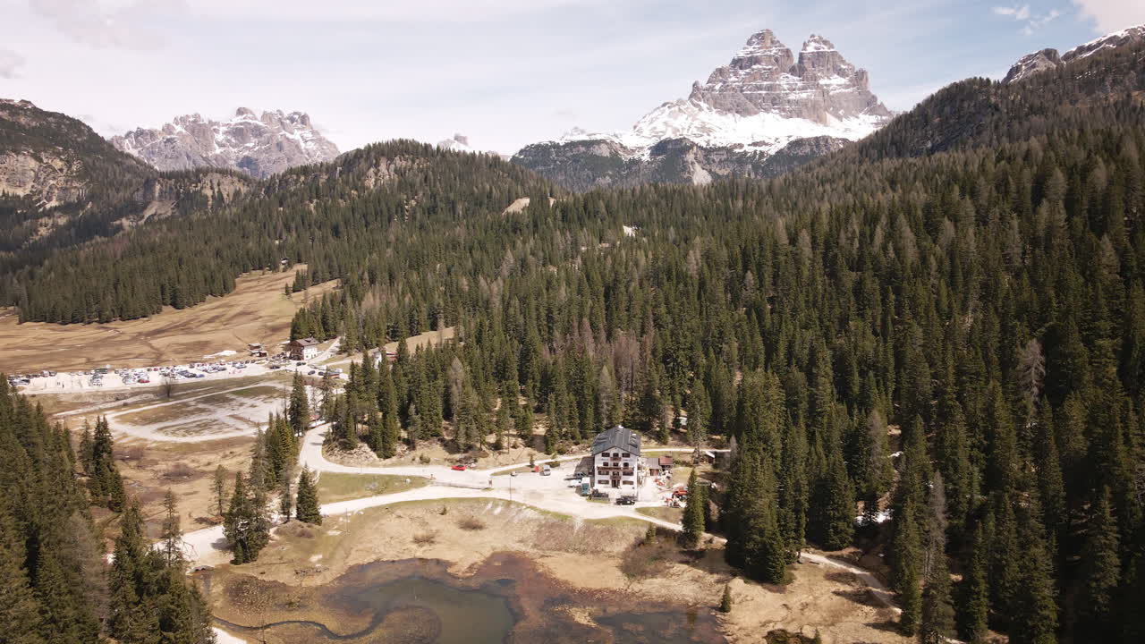 Camera ascends from a small mountain lodge and marshy meadow, revealing dense evergreen forest and towering snow‑capped Dolomite peaks under a bright sky