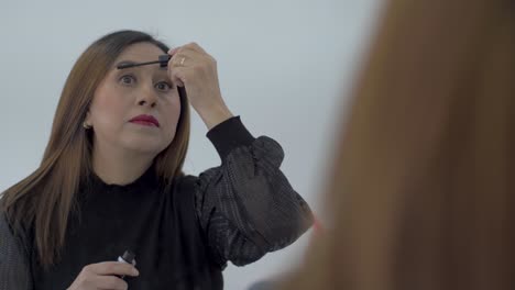 woman with brown hair applying mascara to eyelashes, wearing black long sleeved top using a large mirror