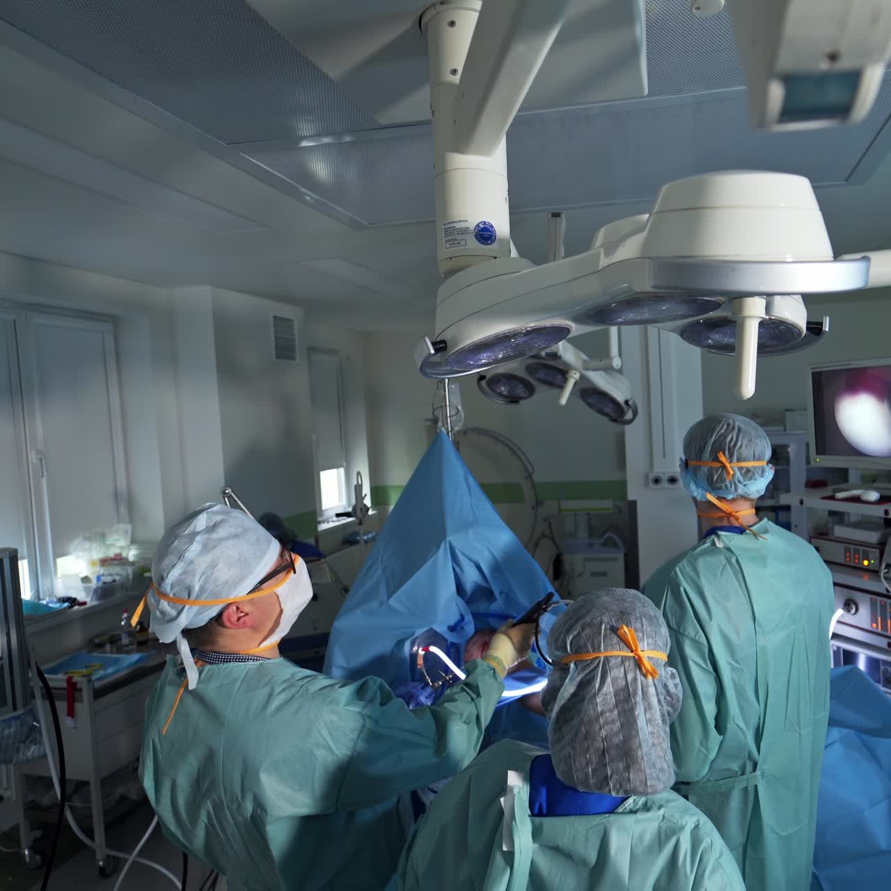 Modern highly-equipped operational room with surgery in progress. Chief surgeon holding the instruments and looks at screen. High angle screen
