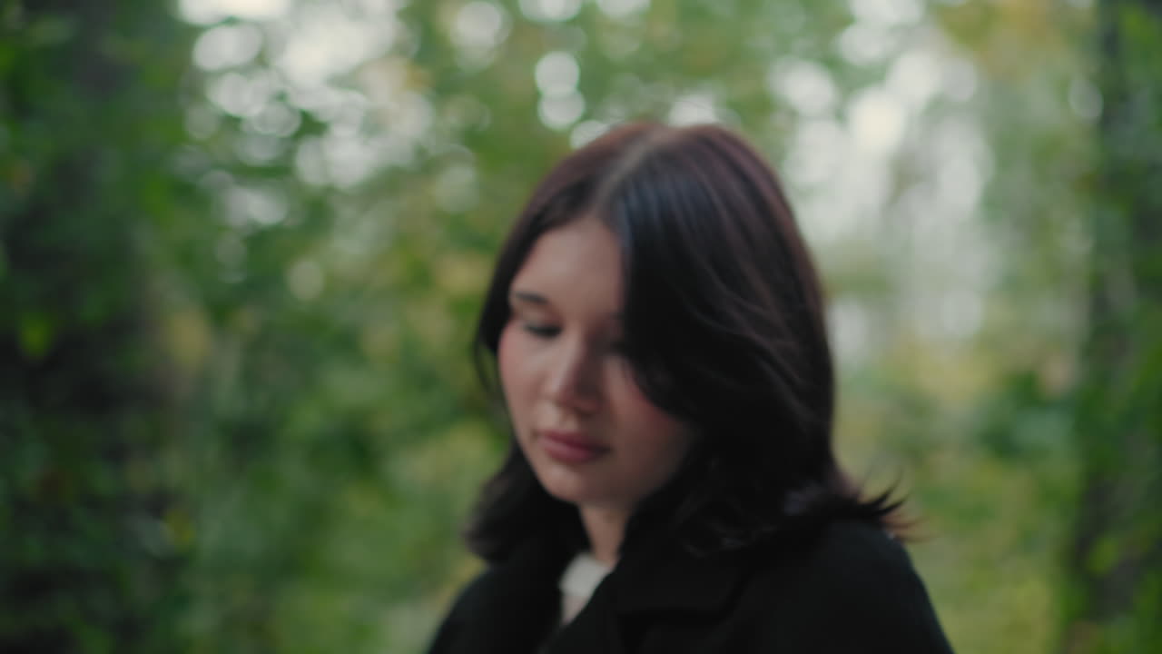 가을 코트를 입은 여성 관광객이 숲길을 거닐면서 책장을 넘기고, 주변을 둘러보며 부드러운 보케 초록빛을 띠고, 차분한 분위기 속에서 산책 중 조용히 독서하며 나뭇잎 아래 평화로운 야외를 만끽해요.