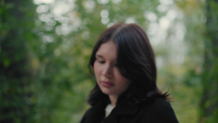 Turista donna con cappotto autunnale che sfoglia le pagine di un libro mentre passeggia su un sentiero nel bosco, guardandosi intorno, verde bokeh delicato, atmosfera calma, pausa di lettura silenziosa durante la passeggiata, fuga pacifica all'aperto sotto le foglie