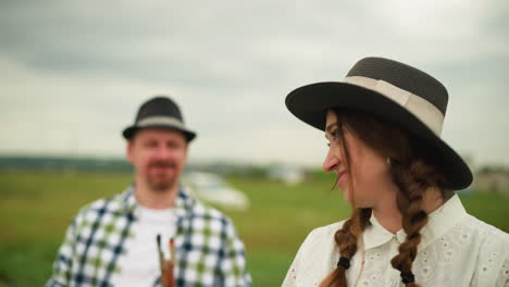 a close-up of a woman in a white dress and a wide-brimmed hat, smiling shyly as she looks to the side. a man in the background, wearing a plaid shirt, points in a direction