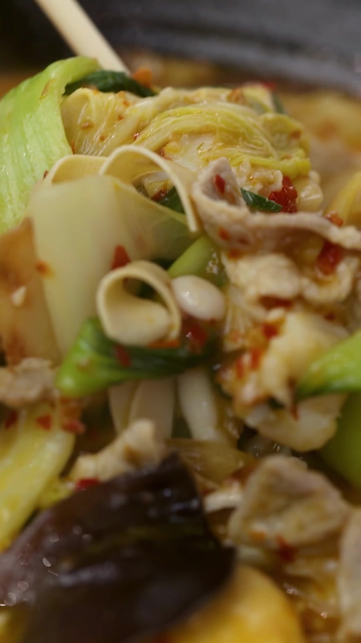 Sumpit close-up mengangkat daging babi, kulit tahu, dan sayuran dari kuah hotpot merah pedas yang mendidih