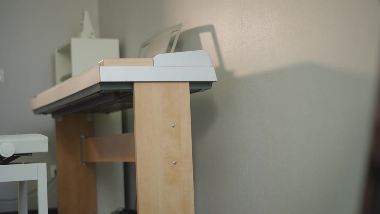 Primo piano sfocato che incornicia la tastiera di un pianoforte, punto di vista di una lezione a casa con profondità di campo ridotta, luce laterale calda e ombra morbida su parete strutturata, leggio in legno per tastiera e sgabello bianco visibile