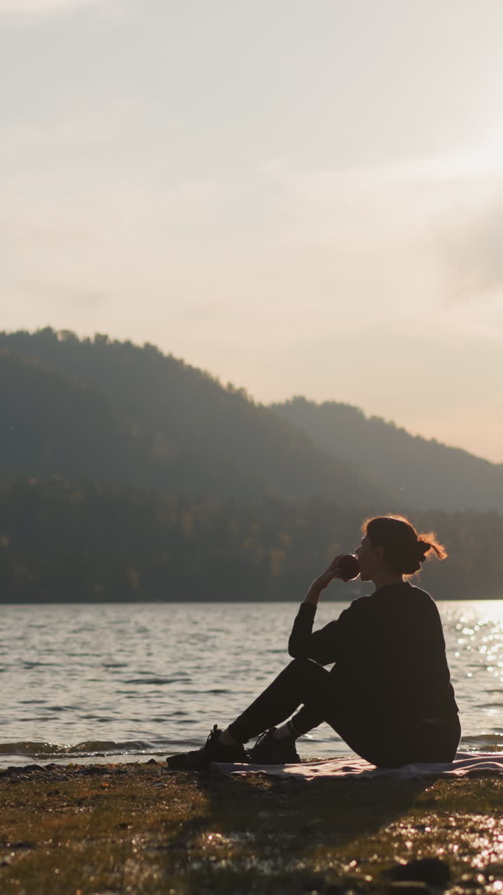 mujer comiendo manzana en un picnic a la orilla del río. mujer sentada junto al río disfrutando de los placeres simples de la vida rodeada de una encantadora vista de las montañas con bosques