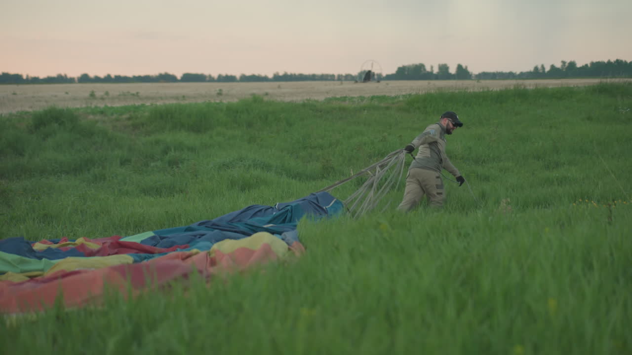 man in cap and gloves unfastening rope from hot air balloon canopy and dragging vibrant colorful fabric across tall grass field at dusk under pastel sky during preflight preparation