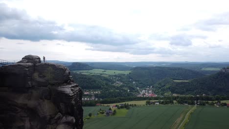 drone passes the summit of an iconic rock formation in europe while man is standing on the mountain top with view of the wide open spaces with clouds