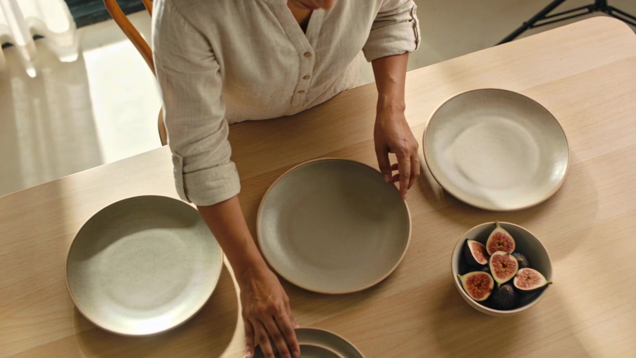 Reaching woman in shirt moving plates on wooden table for styling including fig bowl and tripod