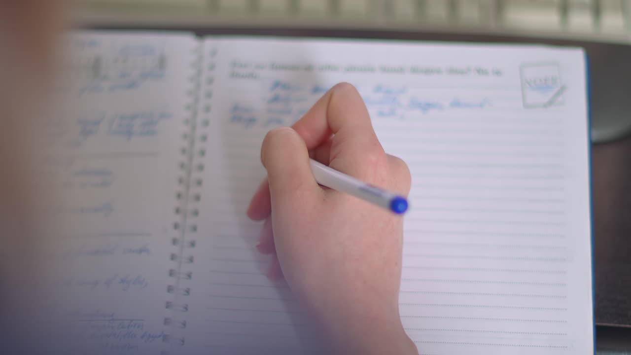 Close up of a woman's hand writing in a spiral notebook with a blue pen while sitting at a desk