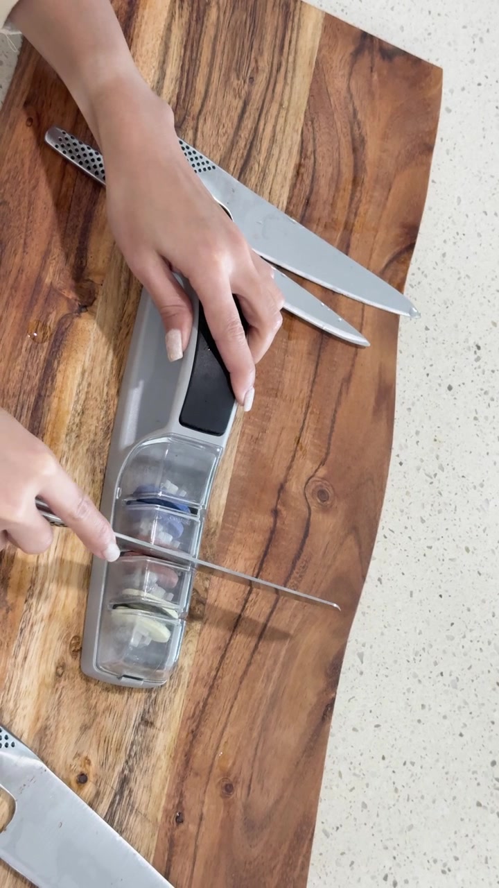 A person sharpens a kitchen knife using a handheld sharpening device on a wooden cutting board in a bright, modern kitchen with overhead lighting