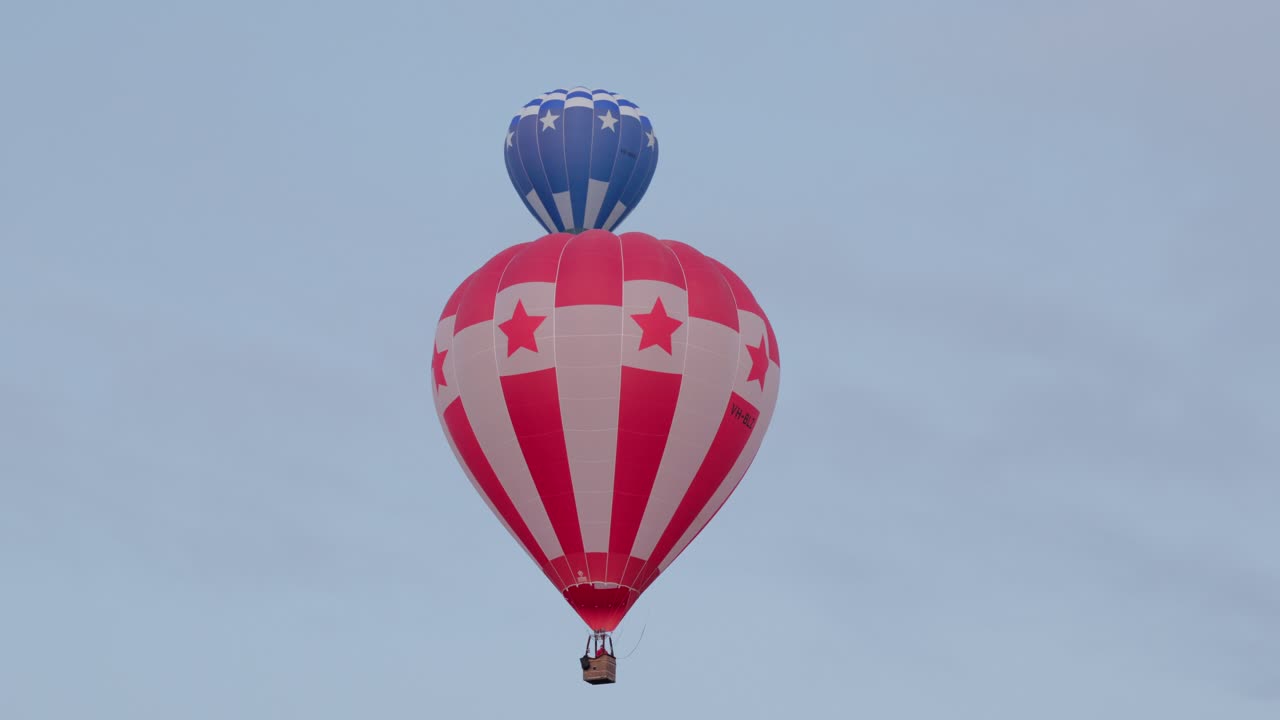 A dynamic scene as one hot air balloon slowly moves, revealing another balloon behind it in the clear sky at the Canberra Balloon Spectacular.