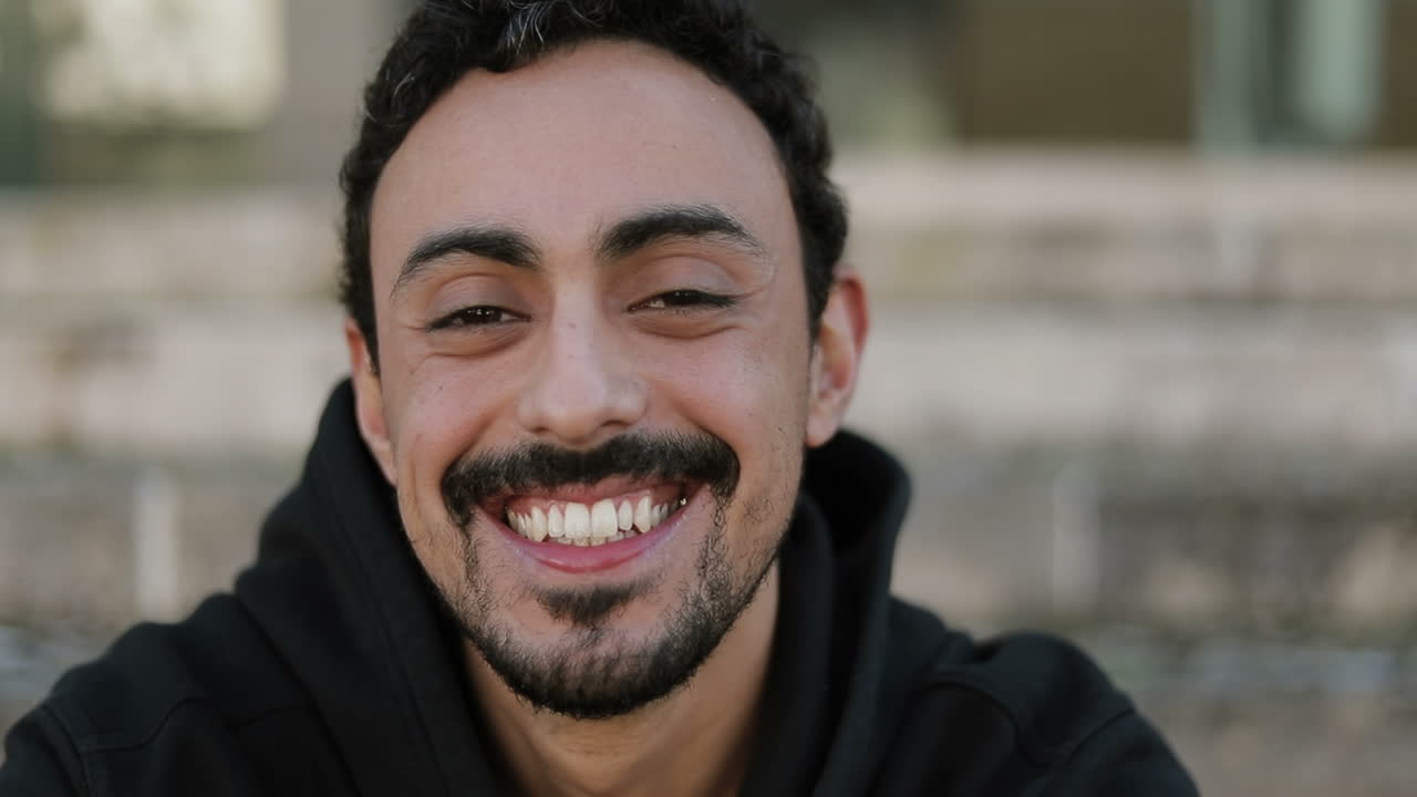 vista frontal de un joven árabe guapo con cabello rizado oscuro y barba con capucha negra sentado en las escaleras afuera