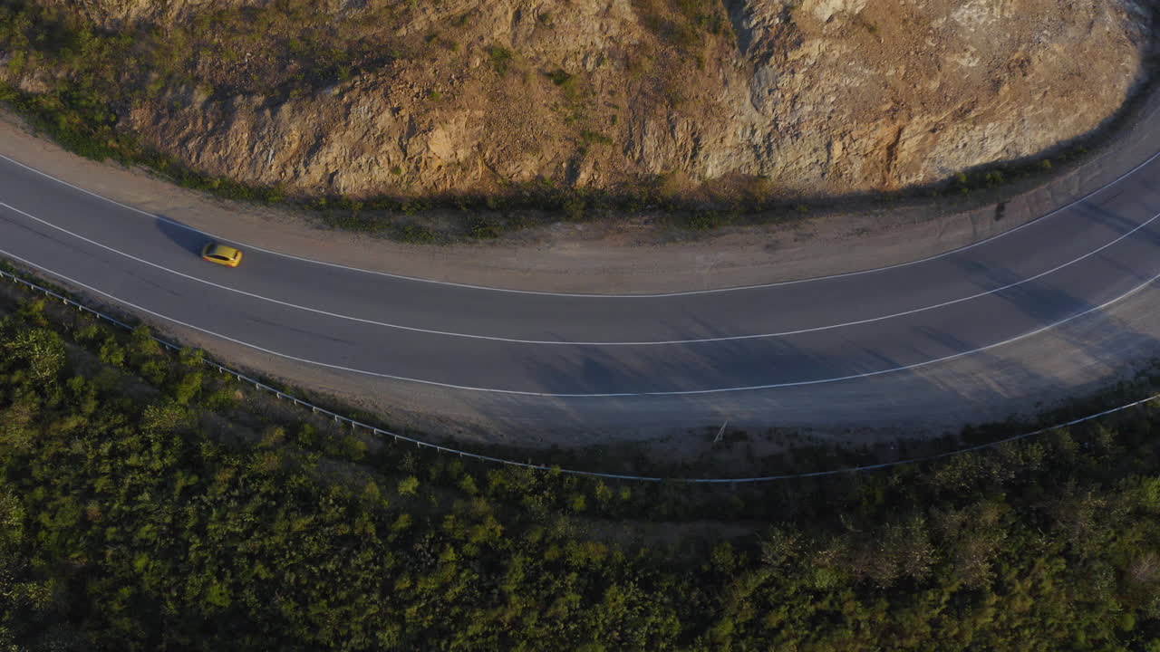 vista superior de un coche de color amarillo que pasa por una larga carretera curva en las montañas verdes con un acantilado en su lado derecho, durante la puesta de sol