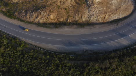 Vista-Superior-De-Un-Coche-De-Color-Amarillo-Que-Pasa-Por-Una-Larga-Carretera-Curva-En-Las-Montañas-Verdes-Con-Un-Acantilado-En-Su-Lado-Derecho,-Durante-La-Puesta-De-Sol