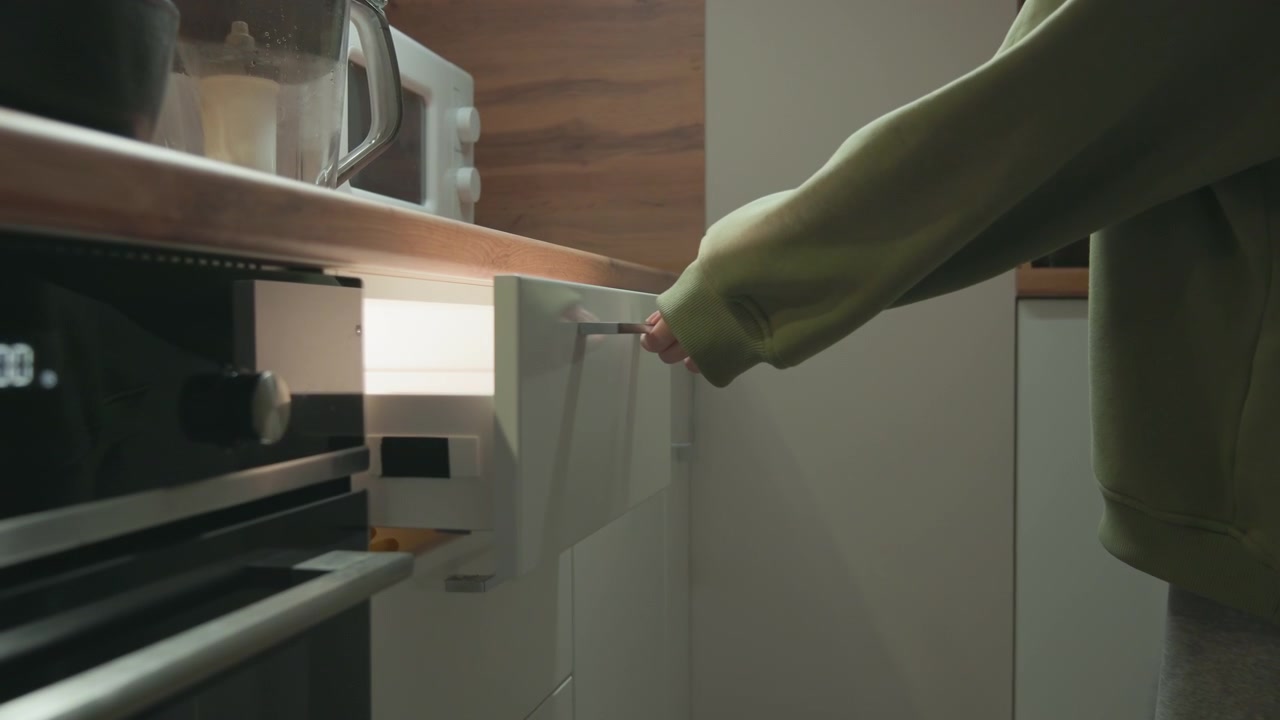 Vista ravvicinata di una mano che si protende in un cassetto della cucina aperto, prendendo un utensile, e poi guidando il cassetto verso l'interno, accanto a mobili bianchi e un piano di lavoro in legno, in un interno domestico moderno con luce diurna soffusa e pulito.