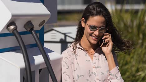 Smiling-caucasian-woman-using-smartphone-by-electric-vehicle-charging-station