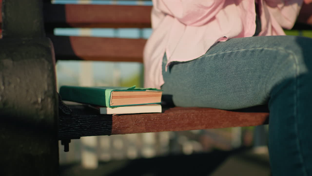 Partial view of individual in jeans and pink top seated on bench with books beside them, outdoor scene with warm sunlight, blurred background featuring iron railing and greenery