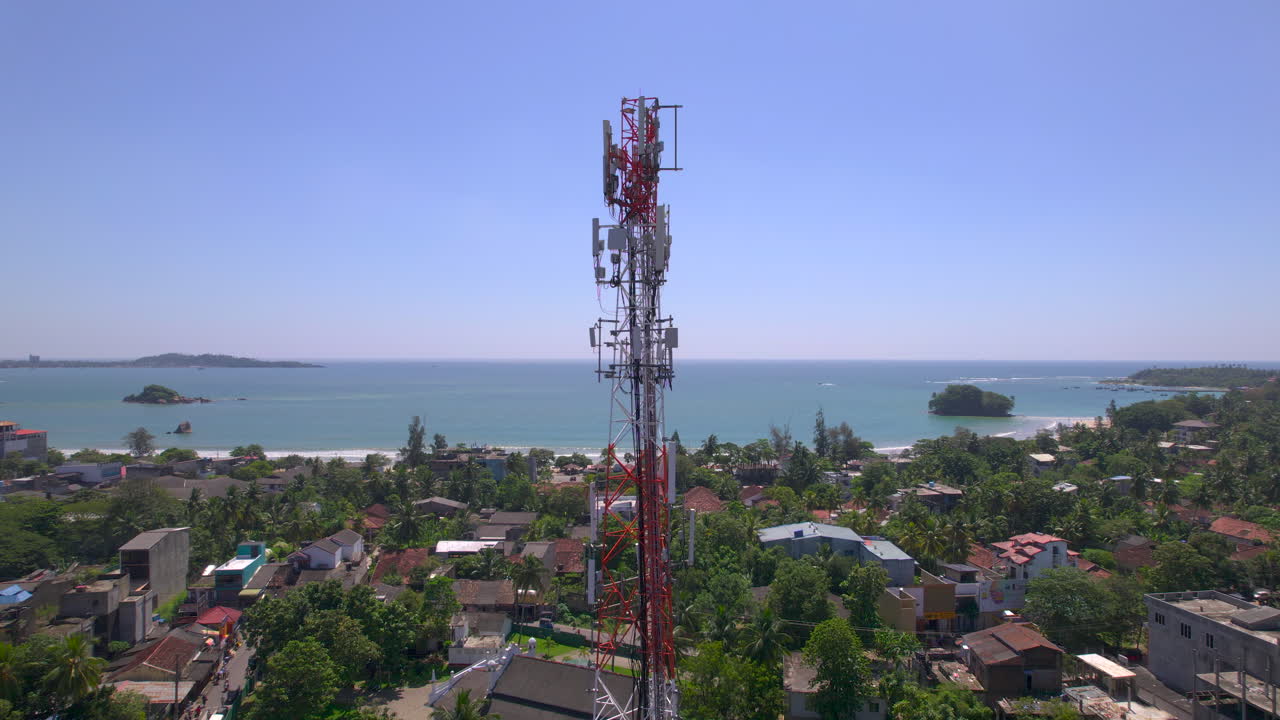 Una torre di telecomunicazioni si erge sopra una città costiera tropicale con tetti di palme e una baia oceanica blu. Infrastruttura di rete mobile in un insediamento marittimo sotto la luce del giorno intensa, sistemi di comunicazione.