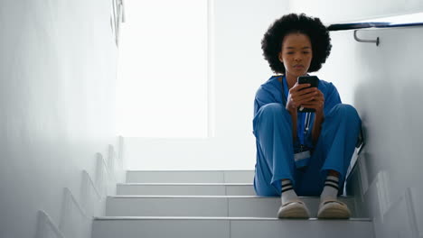 Female-Nurse-Wearing-Scrubs-Browsing-On-Mobile-Phone-Sitting-On-Stairs-In-Hospital