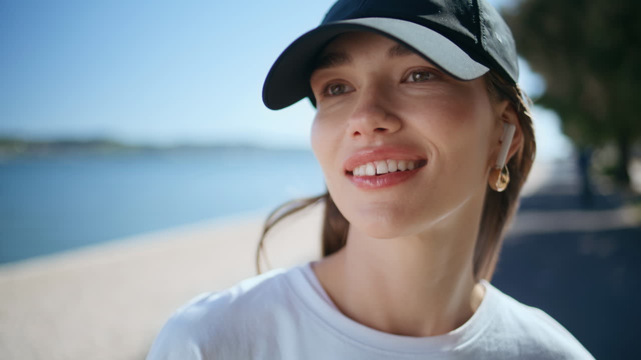 Portrait smiling woman cap standing by riverside summer morning. Girl in earbuds