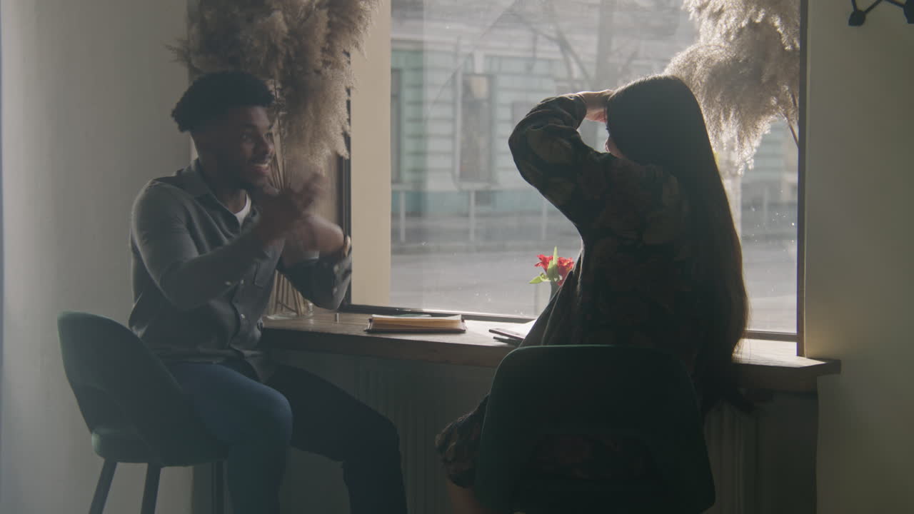 Multi Ethnic Couple Sits at the Table Near Window African American Man Sits at the Table Near Window Gesticulates and Shares Stories with Woman Multi Ethnic Couple on a Date in Cafe Guests in Restaurant with Modern Design Concept of Public Eating