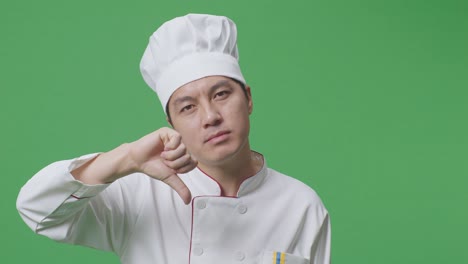 primer plano del chef asiático mostrando los pulgares hacia abajo gesto a la cámara mientras está de pie en la pantalla verde estudio de fondo