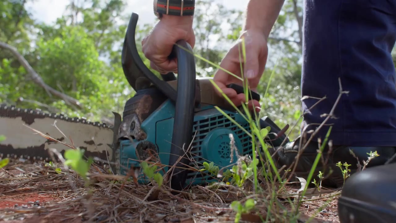 Low-angle close-up of man repeatedly attempting to start an old chainsaw. After several unsuccessful pulls, the starter cord snaps and comes free from the machine, while his child watches