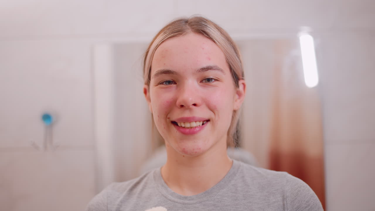 Portrait view of young woman standing in bathroom, looking directly at camera and smiling softly, representing natural beauty, selfcare, confidence, wellness, healthy lifestyle, and morning grooming