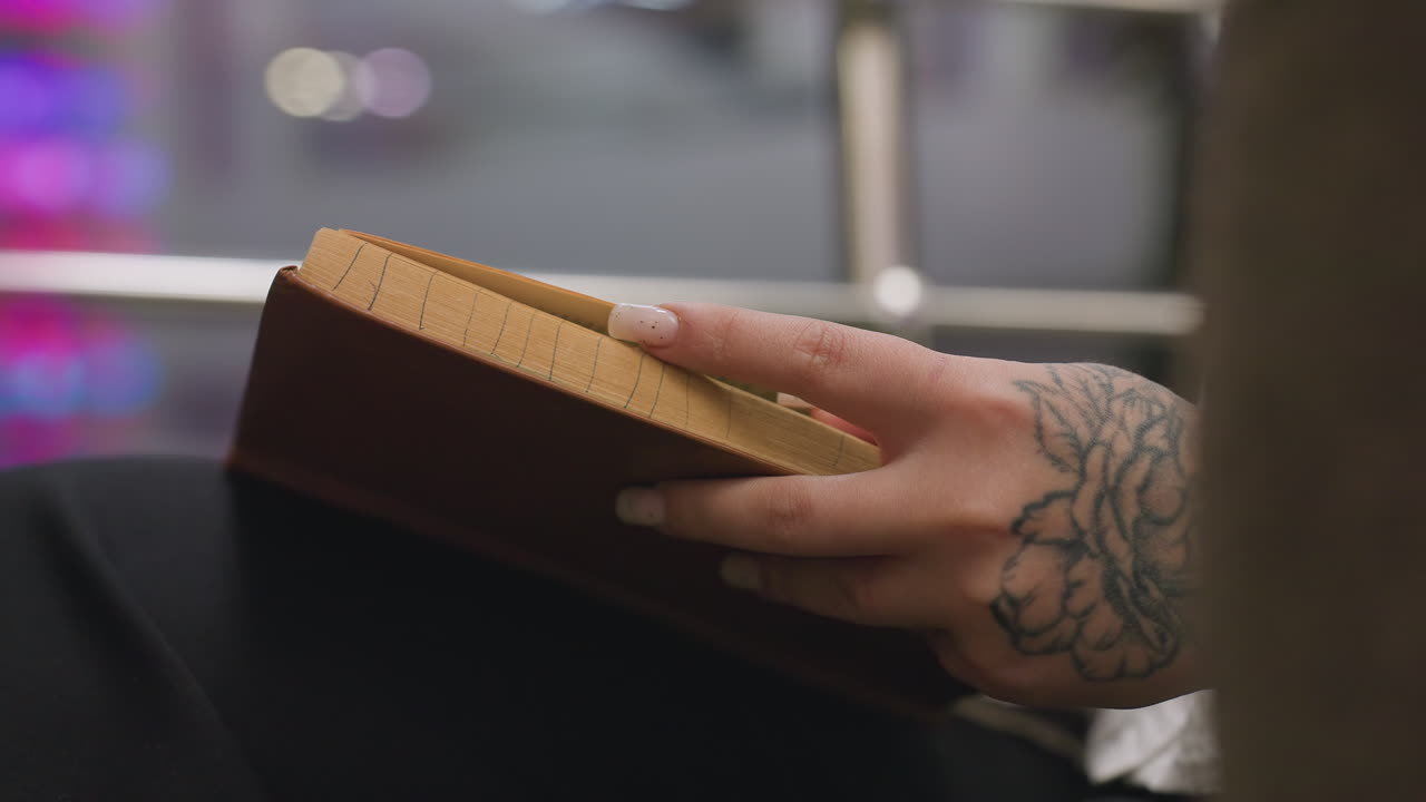 Close up of person with floral tattoo on hand flipping book page while reading indoors, showcasing natural nails and focus on hand movement with blurred indoor background