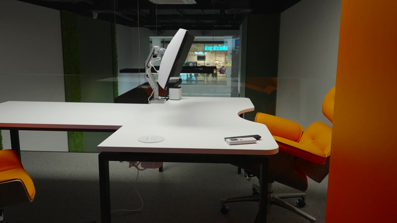 Dimly lit glass-walled conference space with white tables, orange seating, and a wall-mounted monitor in a clean contemporary workplace interior