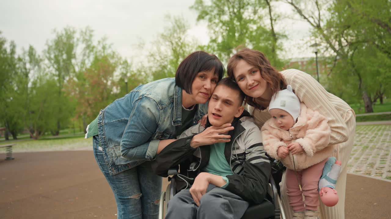 White Family Gathered Around Wheelchair Son In Park, Three Women And Baby Close In Affectionate Group Pose Inclusive Support Network, Gentle Embrace And Hopeful Atmosphere Under Spring Trees