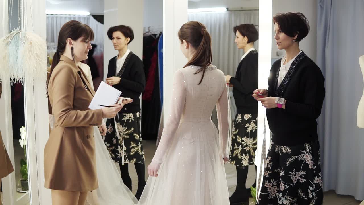 Two female dress designers and a beautiful bride trying her wedding gown in elegant boutique or tailor's studio. A woman in a white dress standing in front the miror. Slow motion