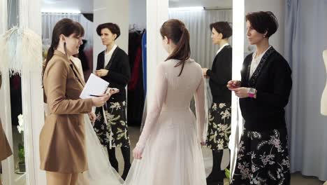 Two female dress designers and a beautiful bride trying her wedding gown in elegant boutique or tailor&#039;s studio. A woman in a white dress standing in front the miror. Slow motion