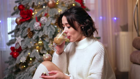 warm drink by the tree. a woman in a cozy sweater sips a warm beverage near a decorated christmas tree, surrounded by soft lights