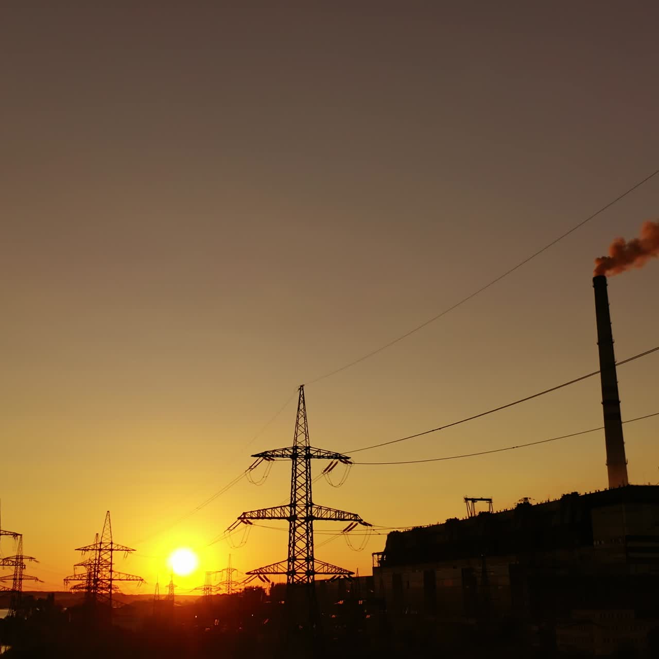 Power station at sunset. Transmission lines near industrial factory with pipes. High voltage electric lines at dusk. Dark smoke from industrial pipes in the evening.