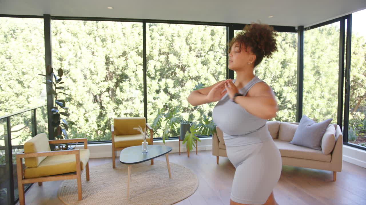 Practicing yoga, plus size woman stretching in living room with large windows