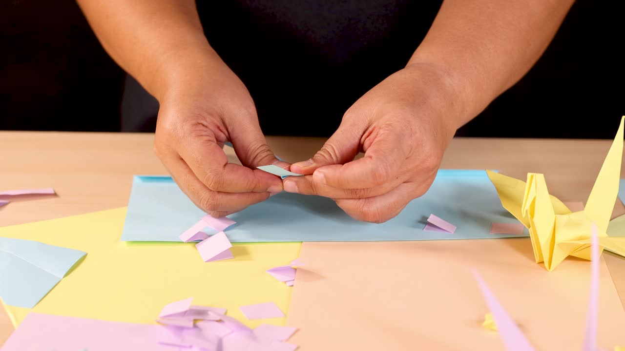 Person folds pastel origami paper with yellow crane nearby, under soft studio lighting, close-up view
