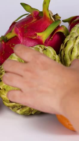la mano tocando una pila de varias frutas tropicales como la fruta del dragón y la cherimoya