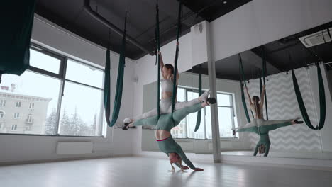 trainer and young woman practising aero gymnastics with suspensions at gym. group of two young beautiful yogi women doing aerial yoga practice in green hammocks in fitness club