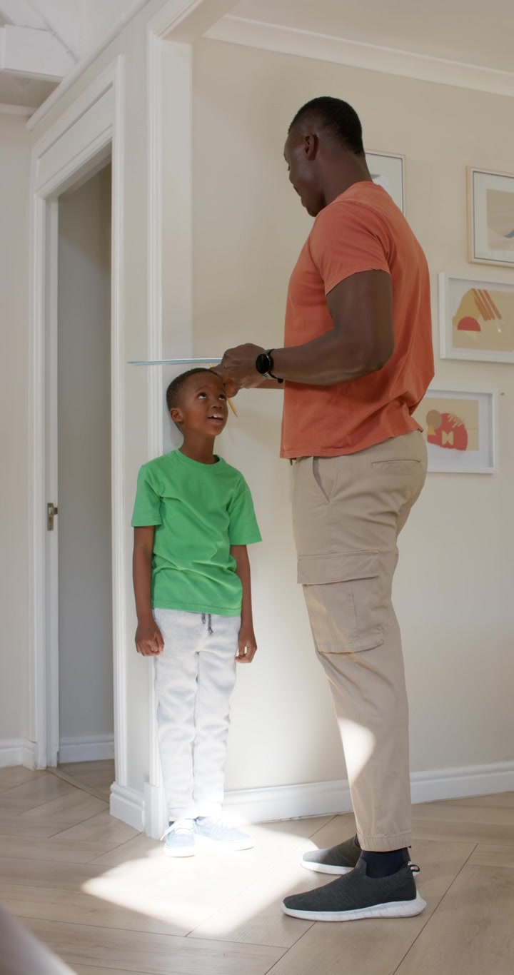 Vertical video: African American father bringing son to painted wall, placing book, marking height