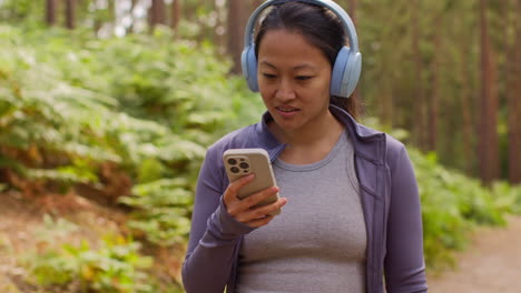 woman wearing wireless headphones exercising doing work out outdoors streaming music or podcast from mobile phone running along track in forest wearing sports clothing 1