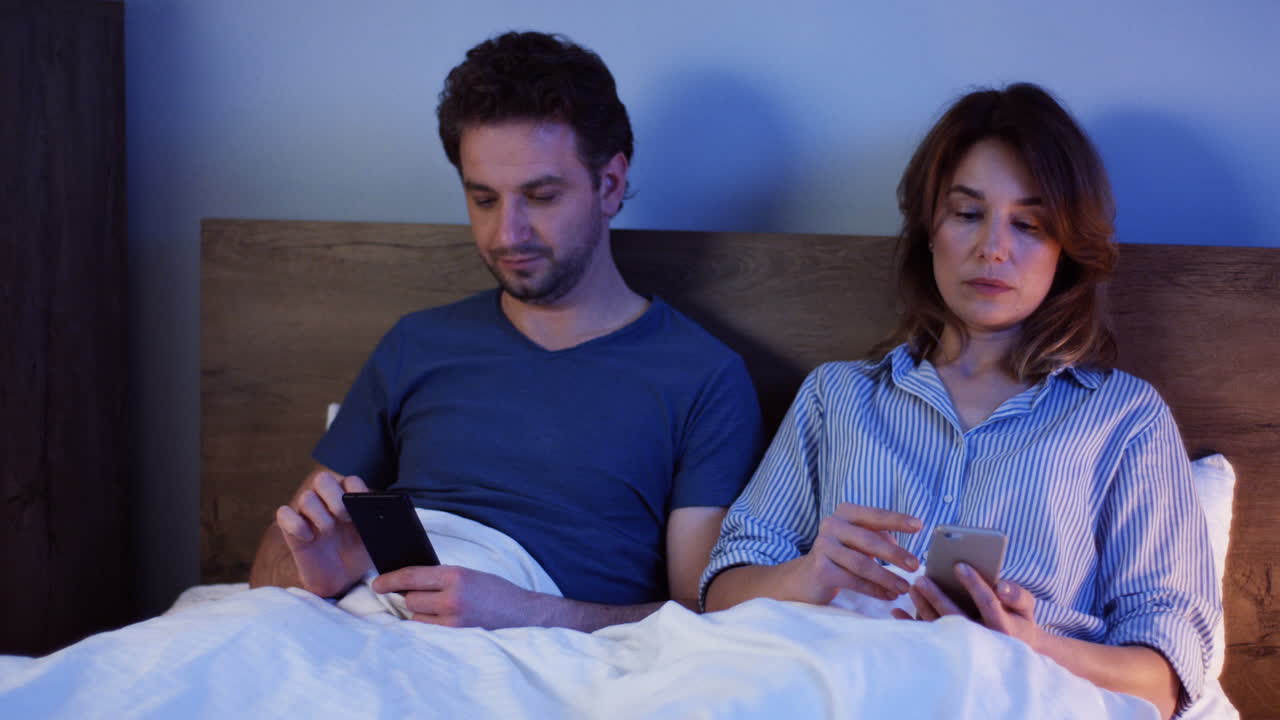 pareja caucásica sentada en la cama y usando smartphones, luego le dicen buenas noches a sus hijos