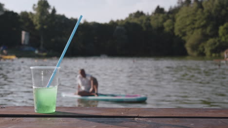 beber en vaso de plástico con paja y persona en paddleboard en segundo plano, verano o vacaciones en el lago