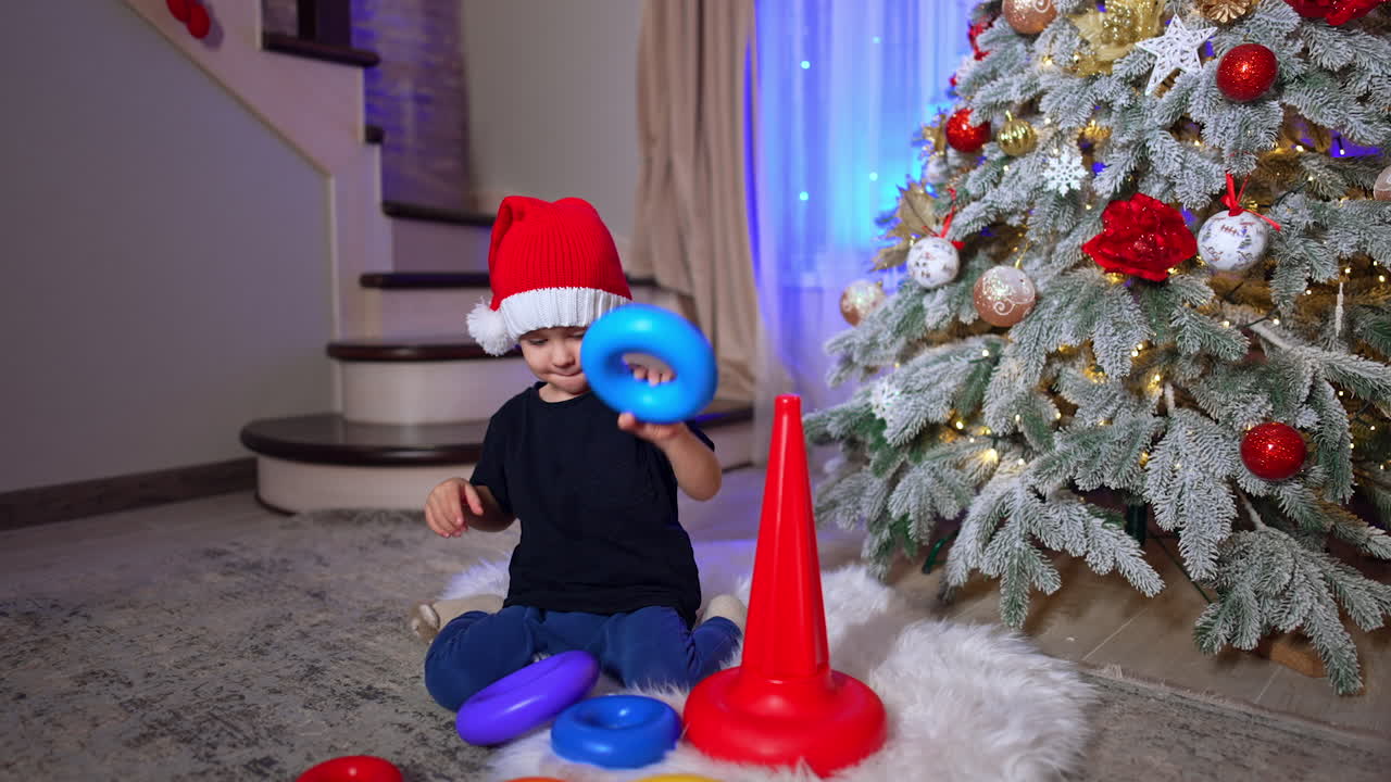 Smart Caucasian toddler wearing Santa cap assembling pyramid. Black cat passes near the kid playing at Christmas tree.